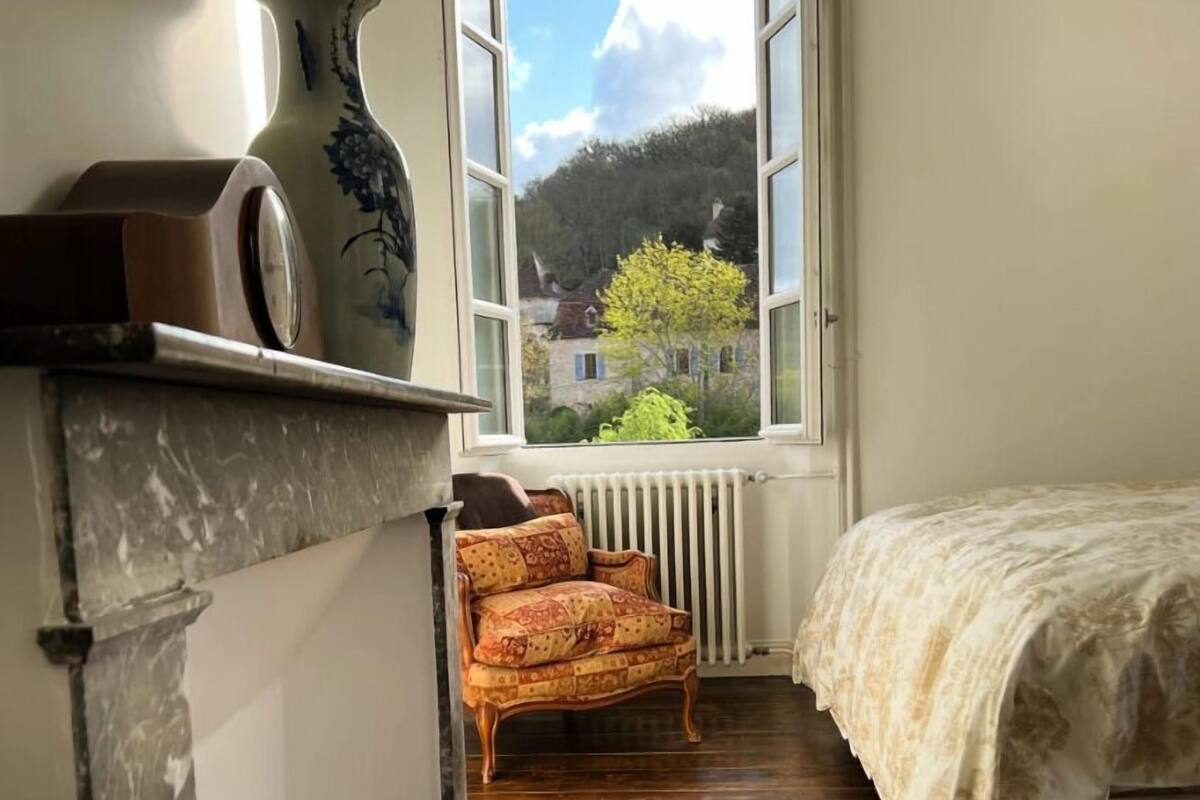 A narrow room with a bed, a radiator, a window with a view of houses and trees, and a patterned armchair.