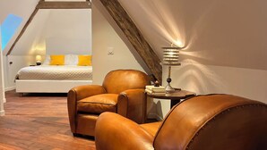 A bedroom with a sloped ceiling, wooden beams, a bed with yellow pillows, a brown leather armchair, and a bedside table with a lamp. - Noubar  (Cénevières)