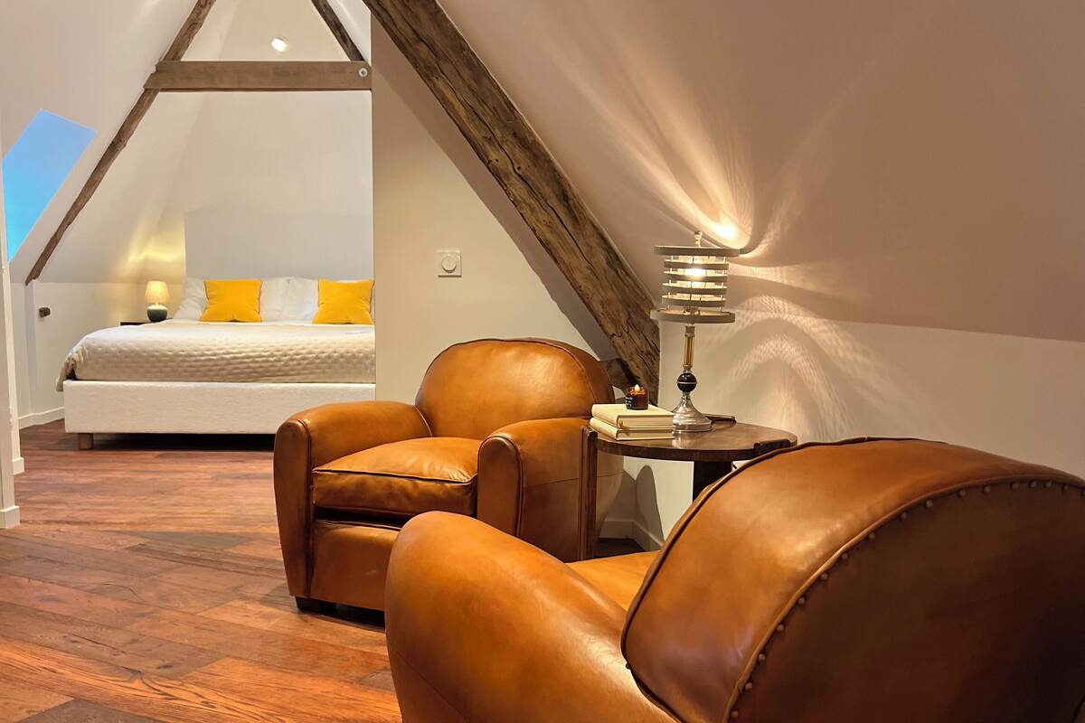 A bedroom with a sloped ceiling, wooden beams, a bed with yellow pillows, a brown leather armchair, and a bedside table with a lamp.