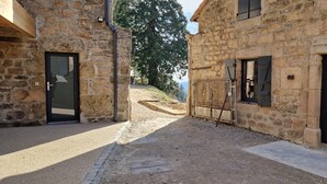 Un bâtiment en pierre doté de deux portes, dont l’une est surmontée d’un portail en métal, et un chemin de terre qui mène à une colline.