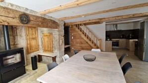 Une cuisine rustique avec un mur en pierre, des poutres en bois, un poêle noir et un coin repas comprenant une table et des chaises.
