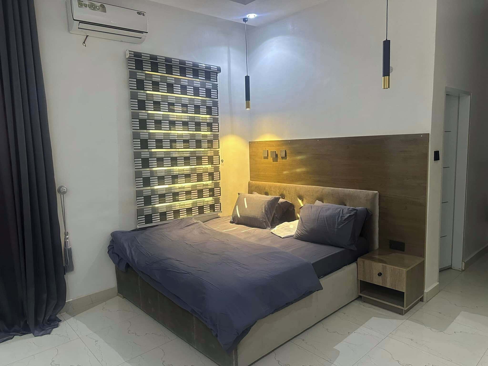 A modern bedroom with a large bed, a headboard with built-in lighting, a wall-mounted air conditioner, and a patterned wall divider.