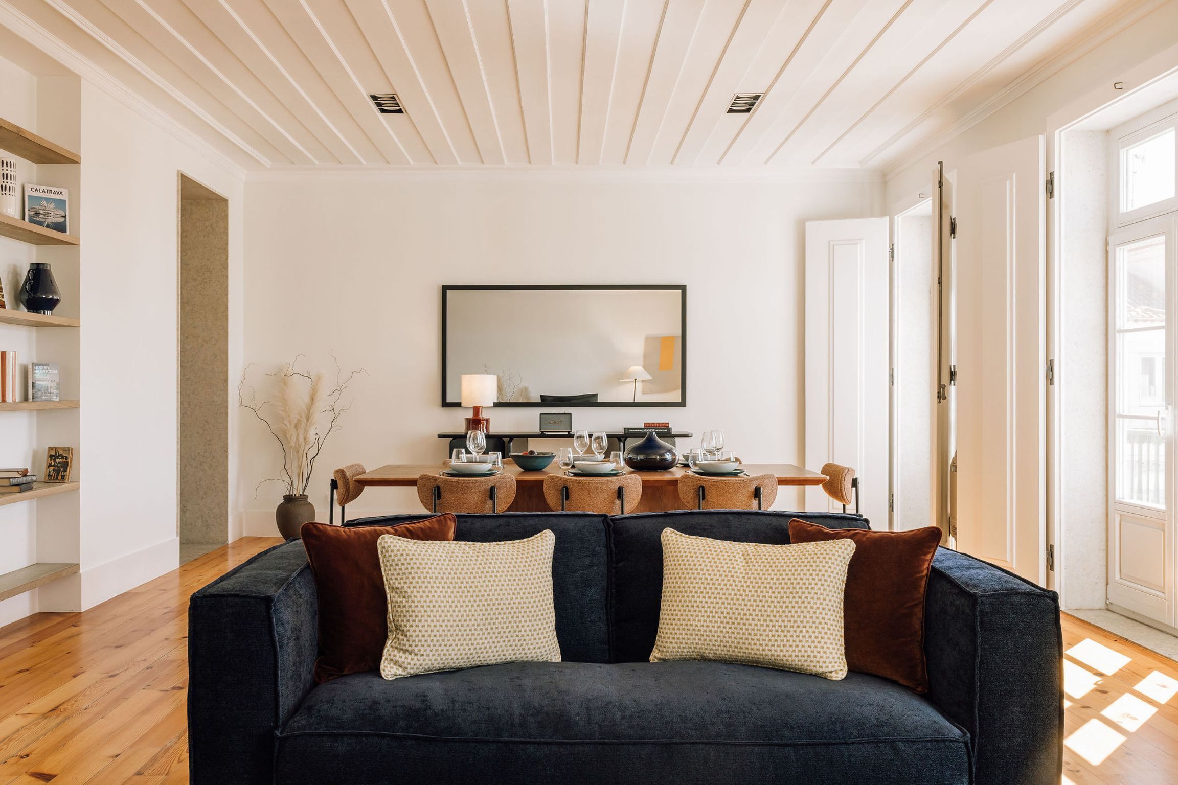 Una sala de estar moderna con un sofá oscuro, una mesa de comedor preparada para cuatro personas y un espejo grande en la pared.