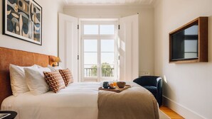 A bedroom with a large bed, a bedside table, a chair, a TV mounted on the wall, and a window with a view of greenery. - Ando Living Corpo Santo House (Lisbon)