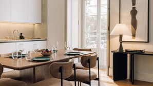 A modern dining area with a wooden table set for six, surrounded by upholstered chairs. A kitchen area with white cabinets and a window is visible in the background. - Ando Living Corpo Santo House (Lisbon)