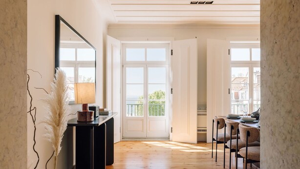 A hallway with wooden flooring, a black console table with a lamp, and a mirror. There are white double doors leading to an outdoor balcony. - Ando Living Corpo Santo House (Lisbon)