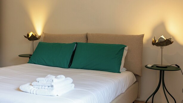A neatly made bed with white linens and green pillows, two bedside tables with lamps, and a stack of white towels. - Vico Santa Maria del Pozzo 9 (Naples)
