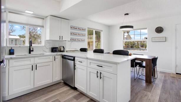 A modern kitchen with white cabinetry, a central island, a dining area with a table and chairs, and a view of the outdoors. - Indian Rocks Beach Gem - Renovated 2BR Beach House (Indian Rocks Beach)