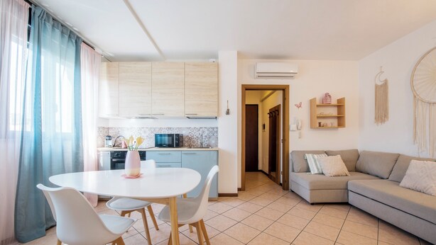 A modern kitchen with light wood cabinets, a white dining table with four chairs, and a grey sofa in the living area.