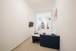 Quarto de hotel estreito com uma cama, uma TV na parede e uma kitchenette ao fundo.