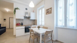 Cocina moderna con armarios blancos, una mesa de comedor y sillas. Se ve una ventana con cortinas translúcidas y una planta en la encimera.