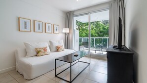 A modern living room with a white sofa, a glass-top coffee table, and a flat-screen TV on a black stand. There are three framed pictures on the wall and a large window with a view of greenery outside. - Ayacucho Apartment | Style & Comfort in Recoleta (Ciudad Autónoma de Buenos Aires)