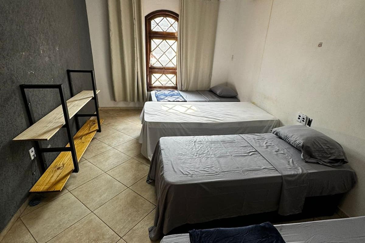 A room with two single beds, a wooden bench, a window with curtains, and an air conditioning unit.