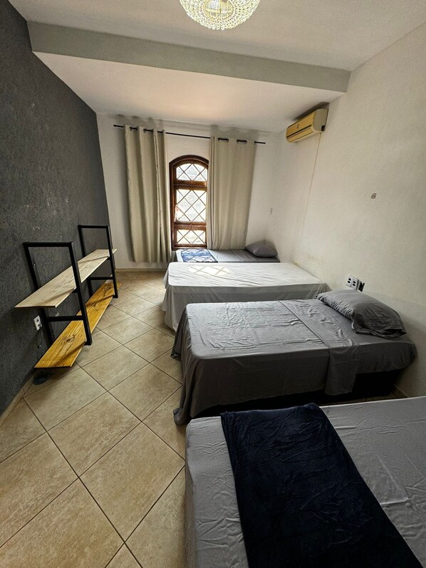 A room with two single beds, a wooden bench, a window with curtains, and an air conditioning unit. - Pousada Castelo da Villa (Foz do Iguacu)