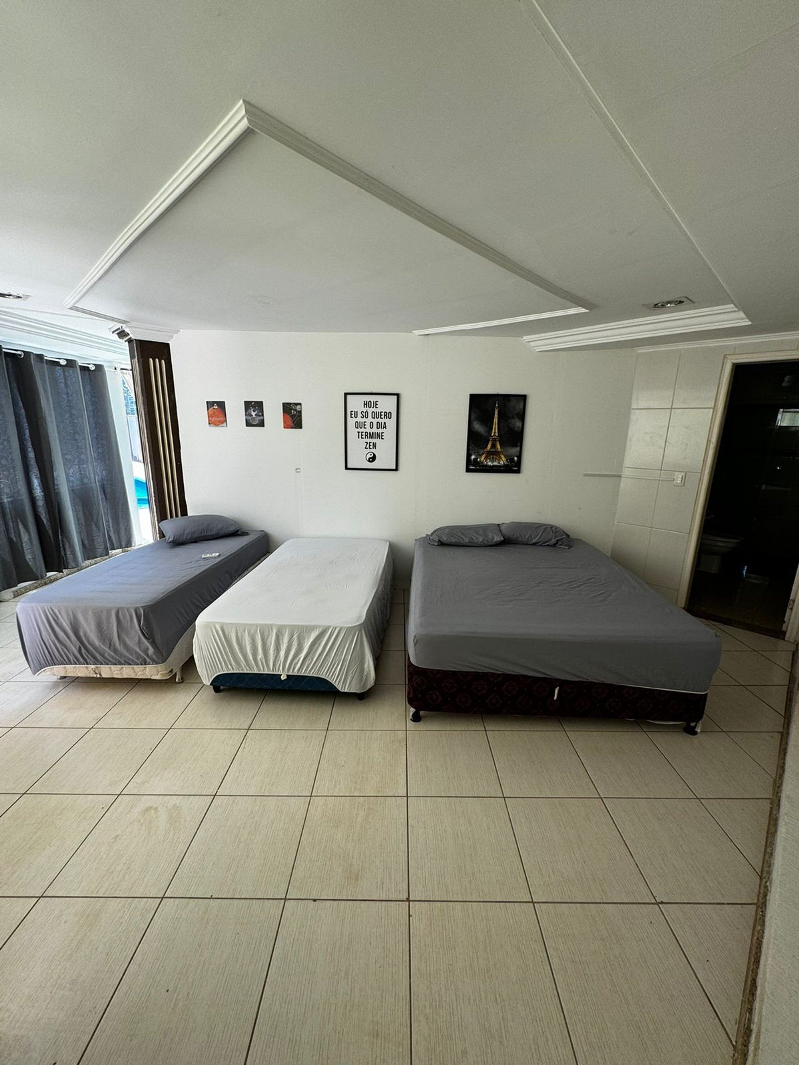 A room with two single beds, a ceiling with recessed lighting, a wall with framed pictures, and a tiled floor.