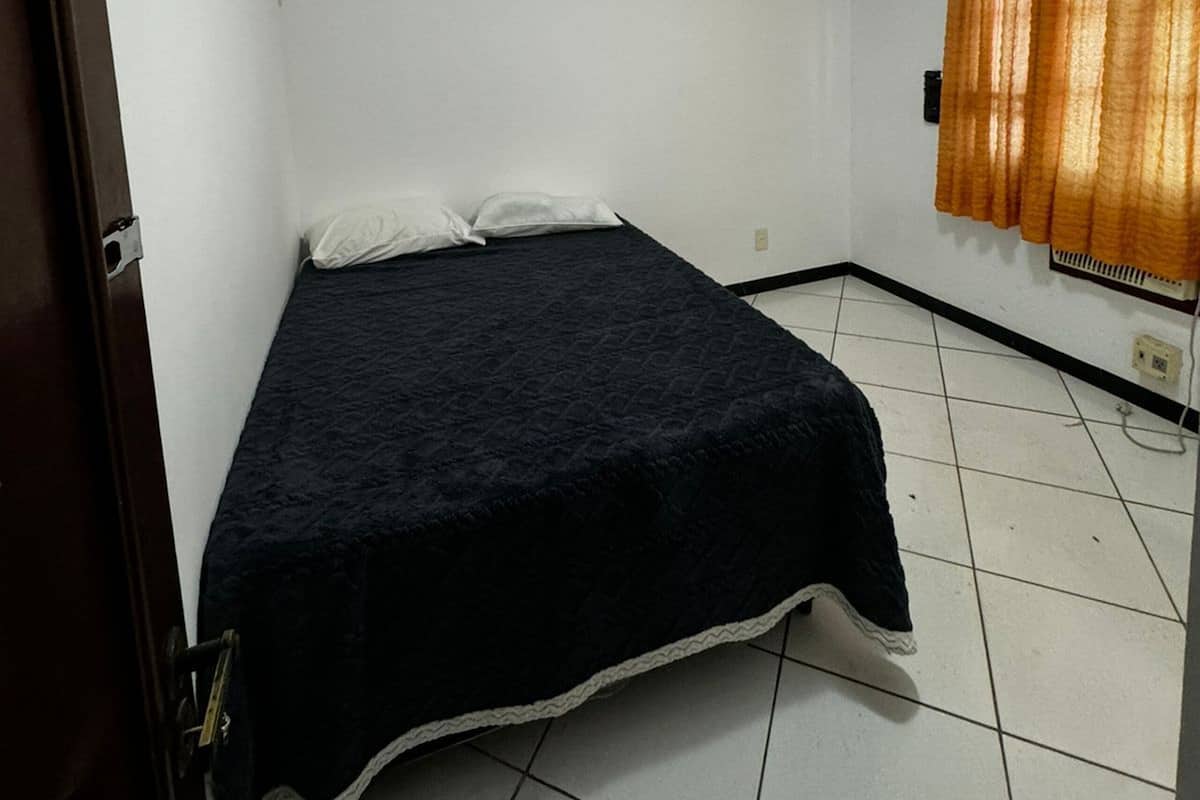 A small, single-bed room with white walls, a tiled floor, a ceiling light, and a window with curtains.