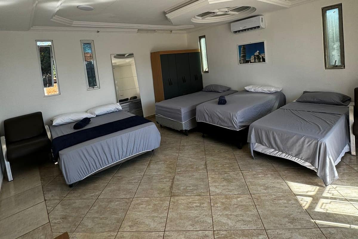 A room with three beds, a desk, and a chair. There are two windows and an air conditioning unit on the wall.