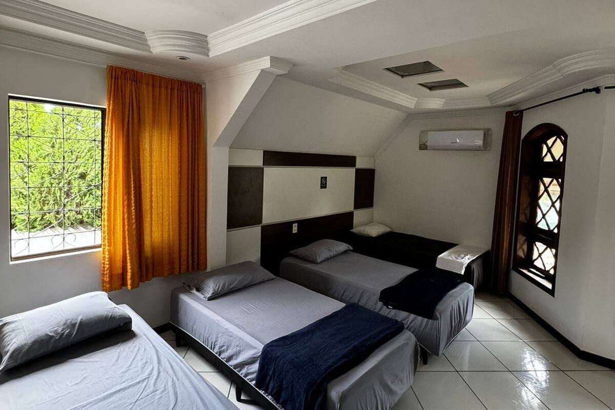 A hotel room with four beds arranged in a U-shape, a large window with curtains, and a view of greenery outside.