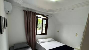 A small, clean room with two single beds, a window with curtains, a ceiling fan, and a white wall with a light switch.