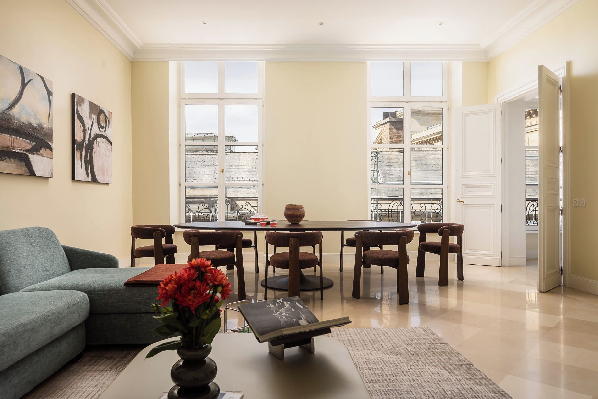 Uma sala de estar com sofá, mesa de centro com um vaso de flores, área de jantar com mesa e cadeiras, e vista através de amplas janelas.