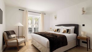 A bedroom with a bed, a chair, a bedside table, a lamp, and a view through a window.