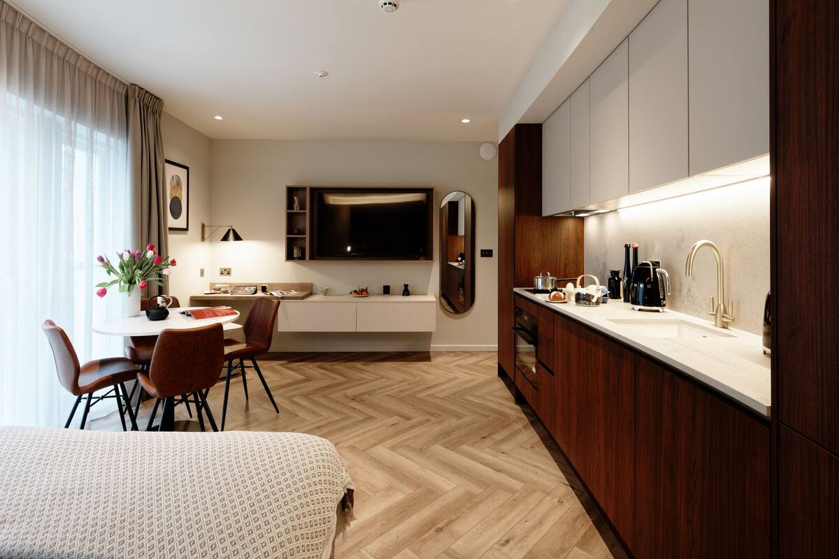 Une chambre d’hôtel moderne dotée d’un coin repas, d’un grand téléviseur à écran plat, de revêtements de sol en bois et d’une cuisine avec un plan de travail blanc.