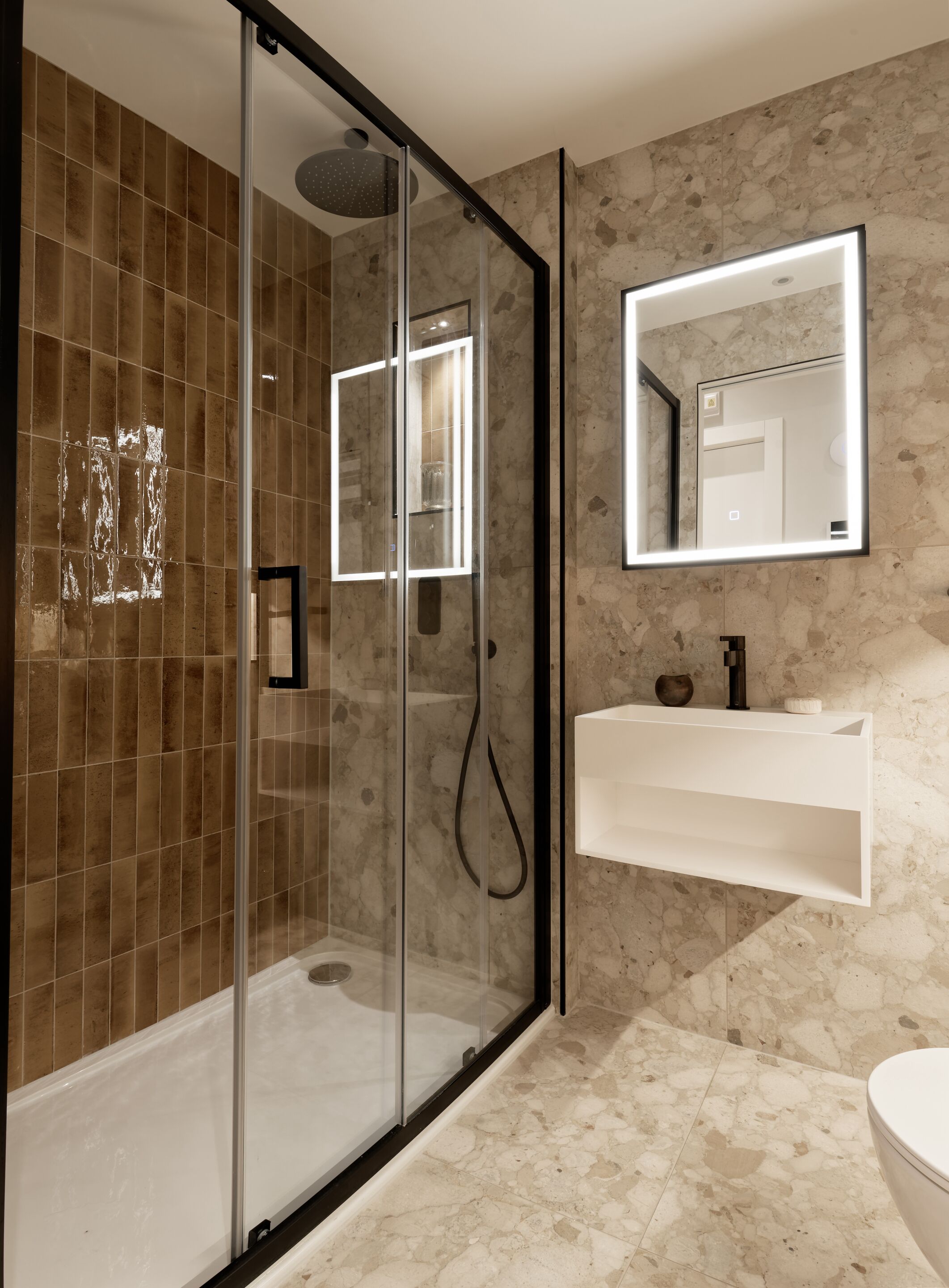 A modern bathroom with a glass-enclosed shower, a marble-tiled wall, a white sink with a mirror above it, and a toilet.