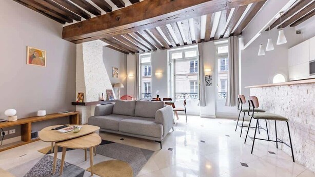 Ein modernes Wohnzimmer mit einer Couch, einem Couchtisch und Sesseln, einem Küchenbereich mit Barhocker und großen Fenstern, die für natürliches Licht sorgen.