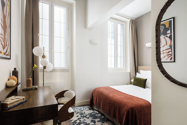 Una habitación de hotel con una cama, un escritorio con una silla, una estantería y dos ventanas.