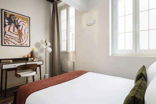 Une chambre d’hôtel avec un lit, un bureau, une chaise, un tableau au mur et une fenêtre.