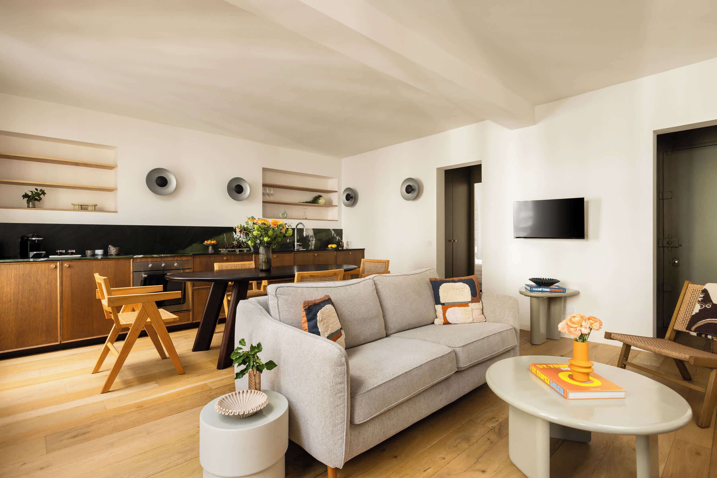 A modern living room with a sofa, coffee table, and chairs. There is a dining area with a table and chairs. A kitchen is visible in the background.