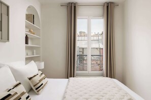 Habitación de hotel con una cama, una estantería, un ventanal con cortinas y vistas a edificios.