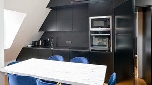 Una cocina compacta con encimera de mármol blanco, dos hornos, un microondas y un refrigerador negro. Hay una mesa de comedor con sillas azules.