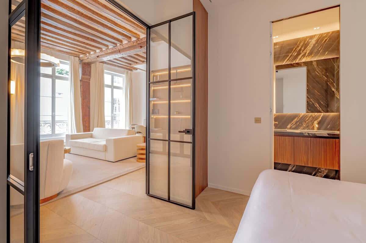A modern hotel room with a bed, a seating area with a white sofa, a glass partition, and a marble vanity.