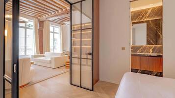 A modern hotel room with a bed, a seating area with a white sofa, a glass partition, and a marble vanity.