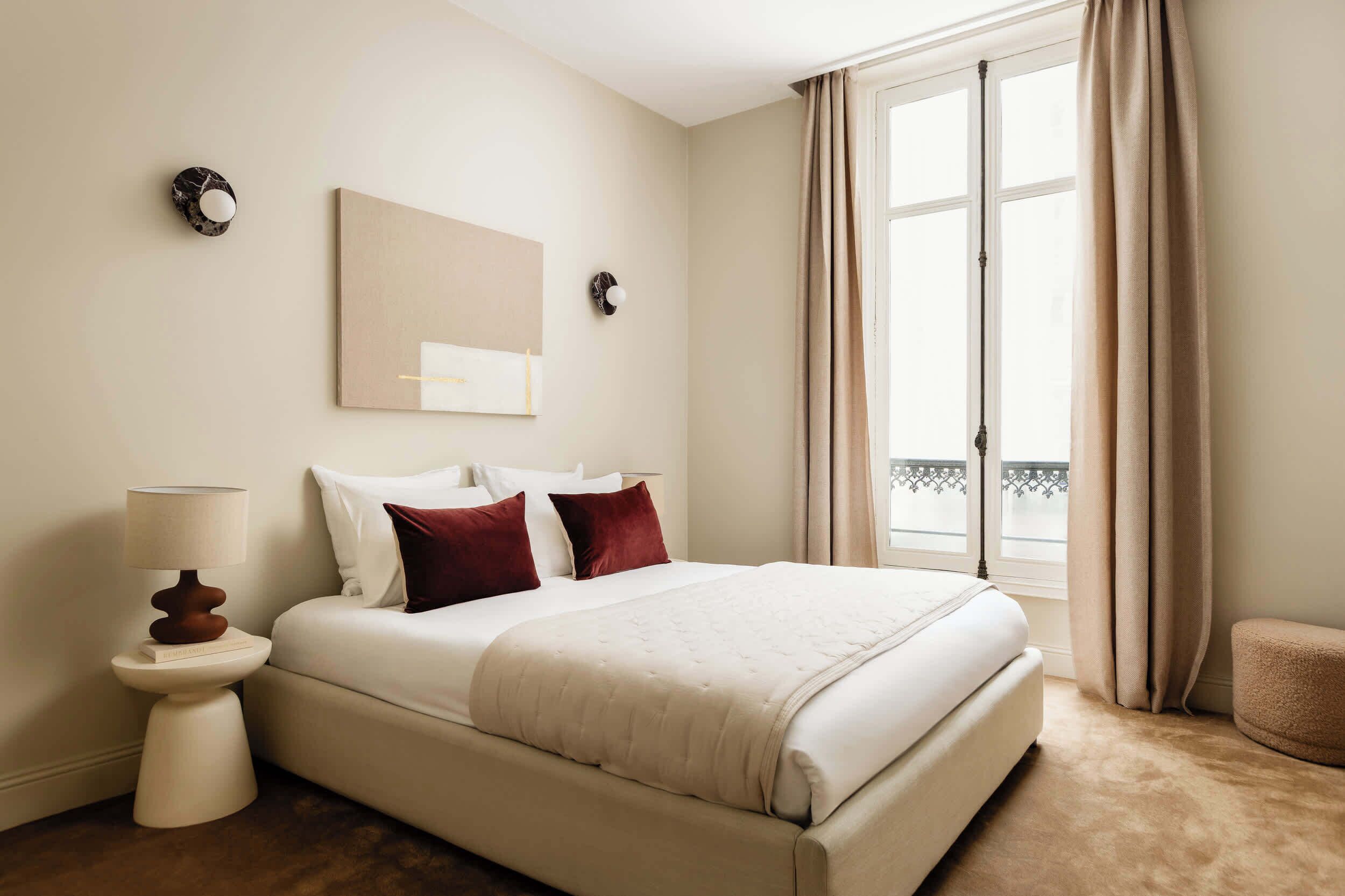 A hotel room with a bed, a bedside table with a lamp, a wall-mounted mirror, and a window with curtains.