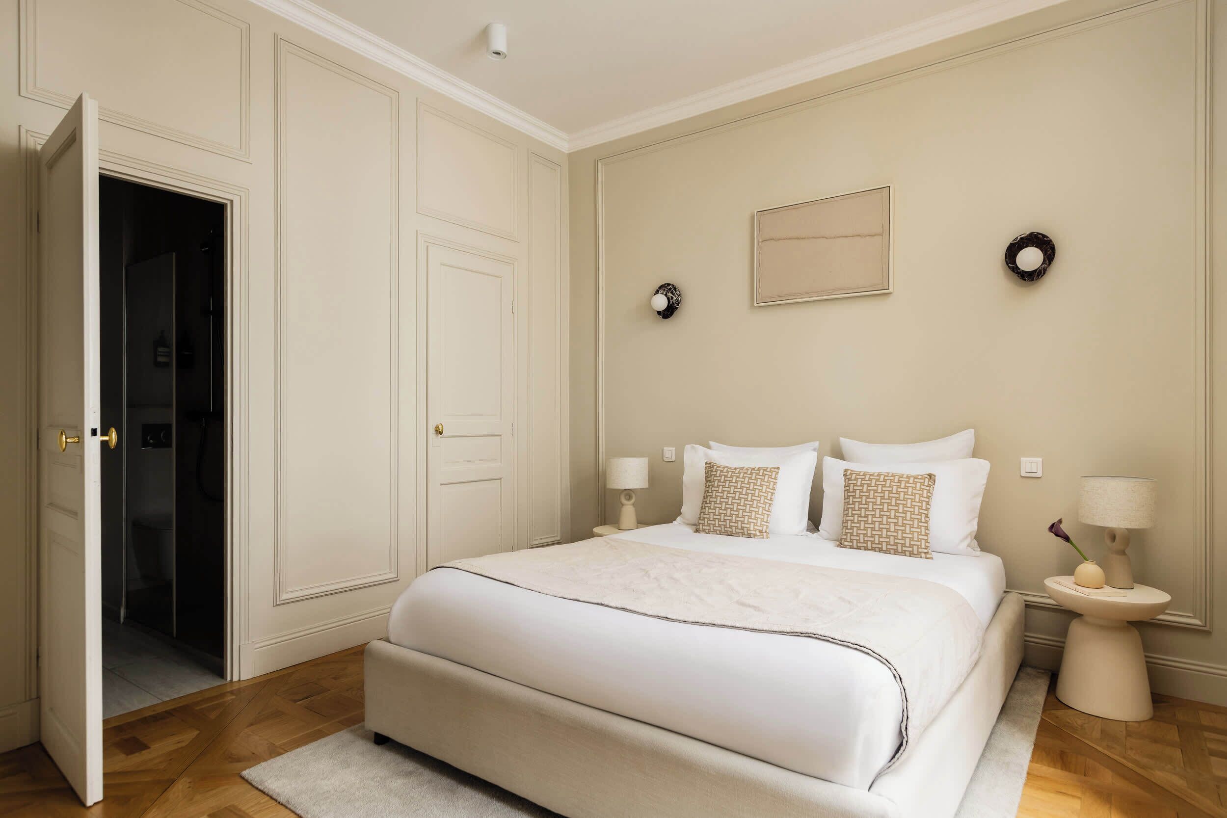 A bedroom with a bed, bedside tables, a door, and a wall-mounted light fixture.