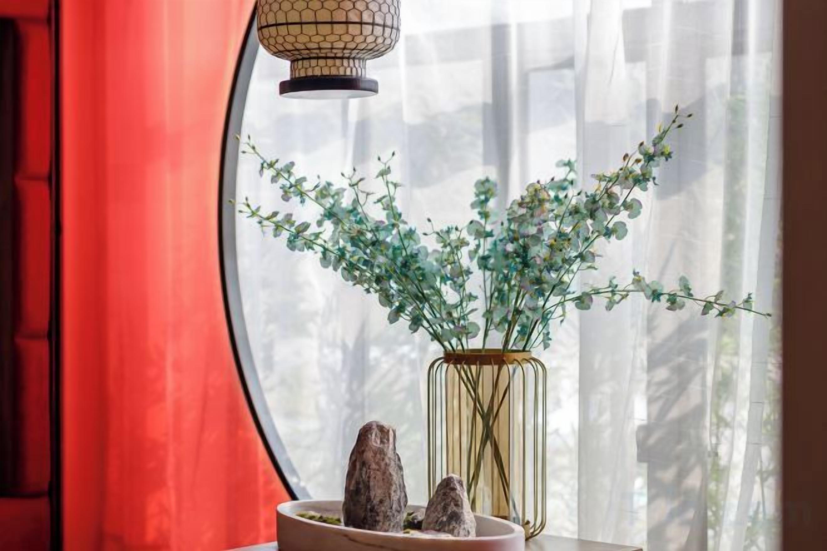 A window with a vase of green plants and two rocks, a red curtain, and a hanging light fixture.