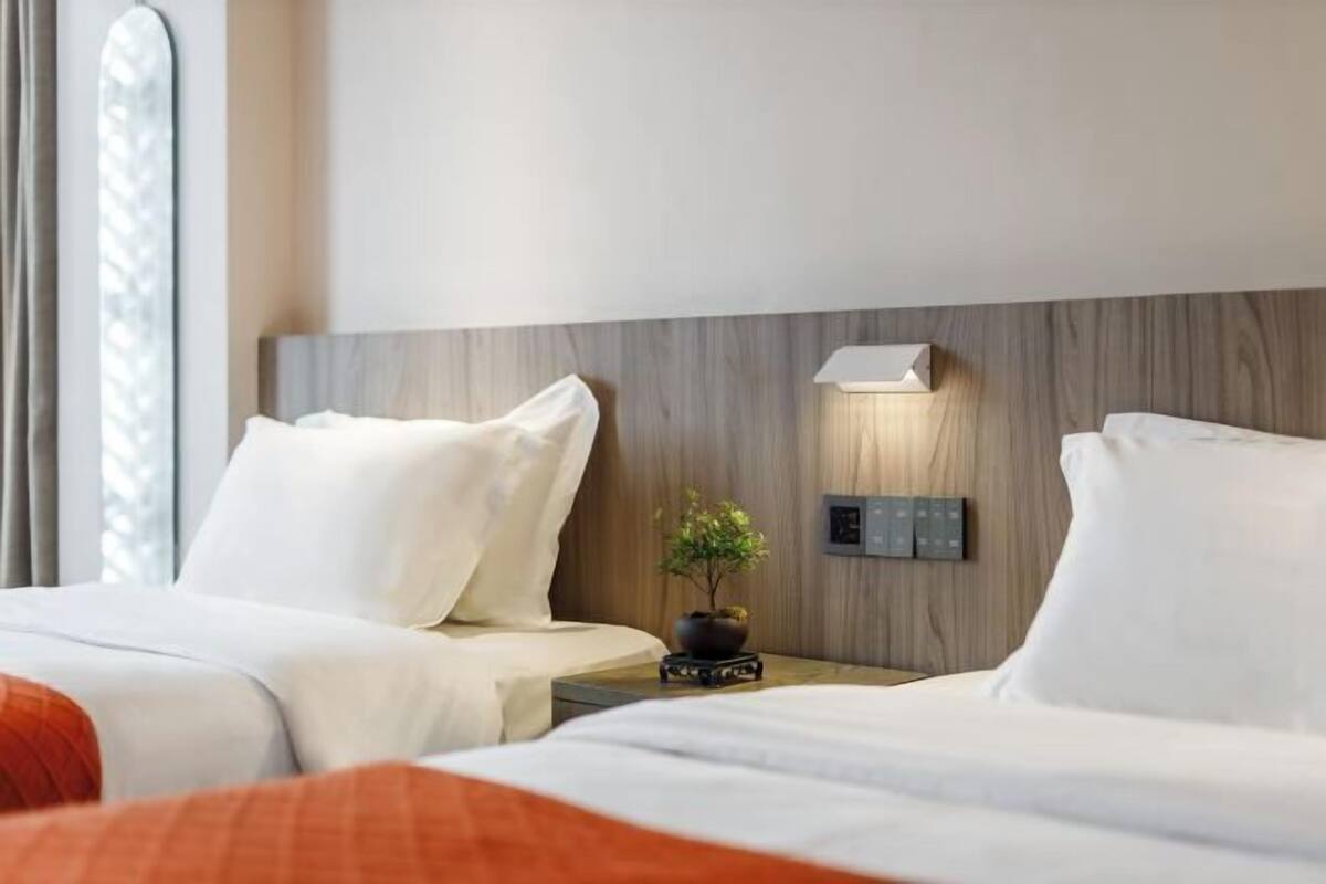 Hotelkamer met twee bedden, een nachtkastje met een lamp, een kleine plant en een bedieningspaneel aan de muur.