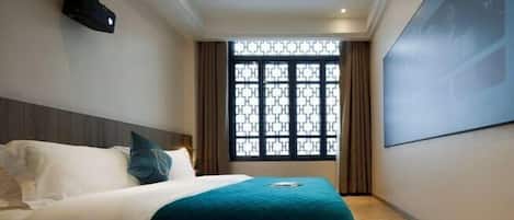 A hotel room with a bed, a projector screen, a window with a geometric pattern, and a wall-mounted light fixture.