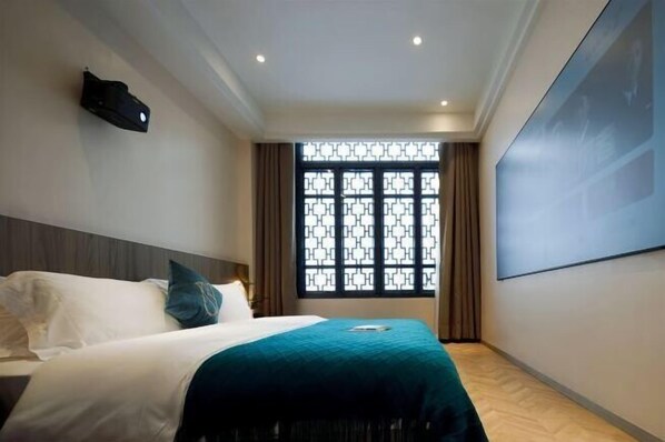 Een hotelkamer met een bed, een projectiescherm, een raam met een geometrisch patroon en een wandlamp.