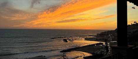 Vista de un atardecer desde un balcón con vista al océano, una costa rocosa y edificios a lo lejos.