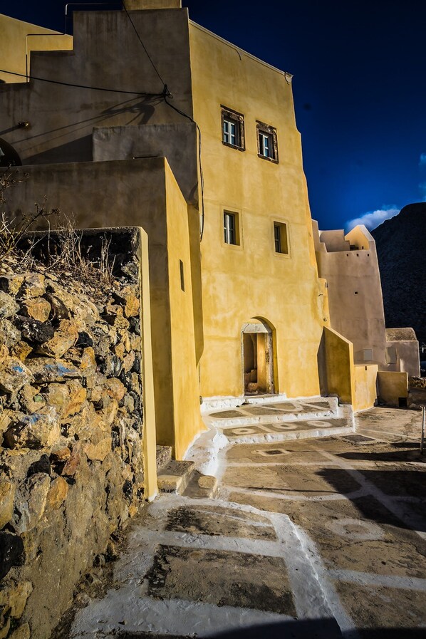 A building with a yellow facade, a stone wall, and a staircase leading to an entrance. - The Commander s Cave (Santorini)