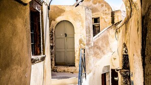 A narrow, arched passageway leading to a staircase with a wooden handrail, flanked by weathered walls and a door at the top. - The Commander s Cave (Santorini)
