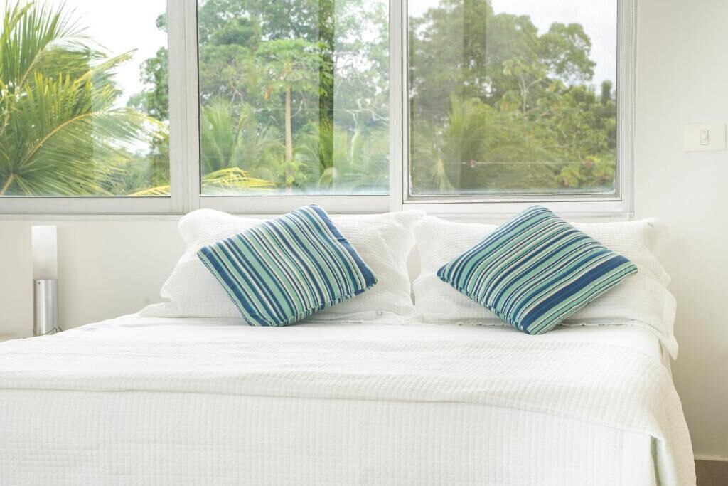 A bed with white bedding and two striped pillows, a window with a view of greenery, and a white wall.