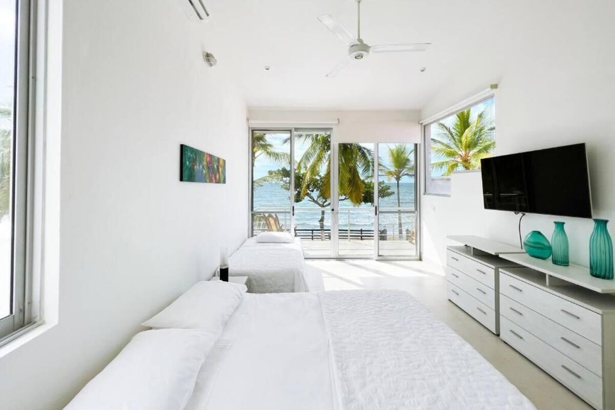 Un dormitorio moderno con una cama grande, un televisor y vistas a palmeras y al mar a través de amplios ventanales.