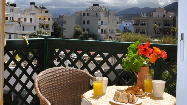 Un balcón con una silla de mimbre, una mesa con mantel, un plato de sándwiches, un jarrón con flores y dos vasos de jugo de naranja.