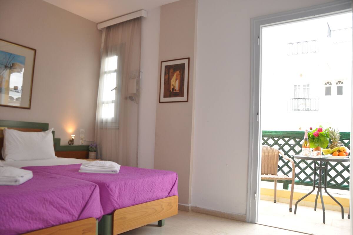 A hotel room with a bed, a nightstand, a framed picture, and a balcony with a table, chair, and fruit.