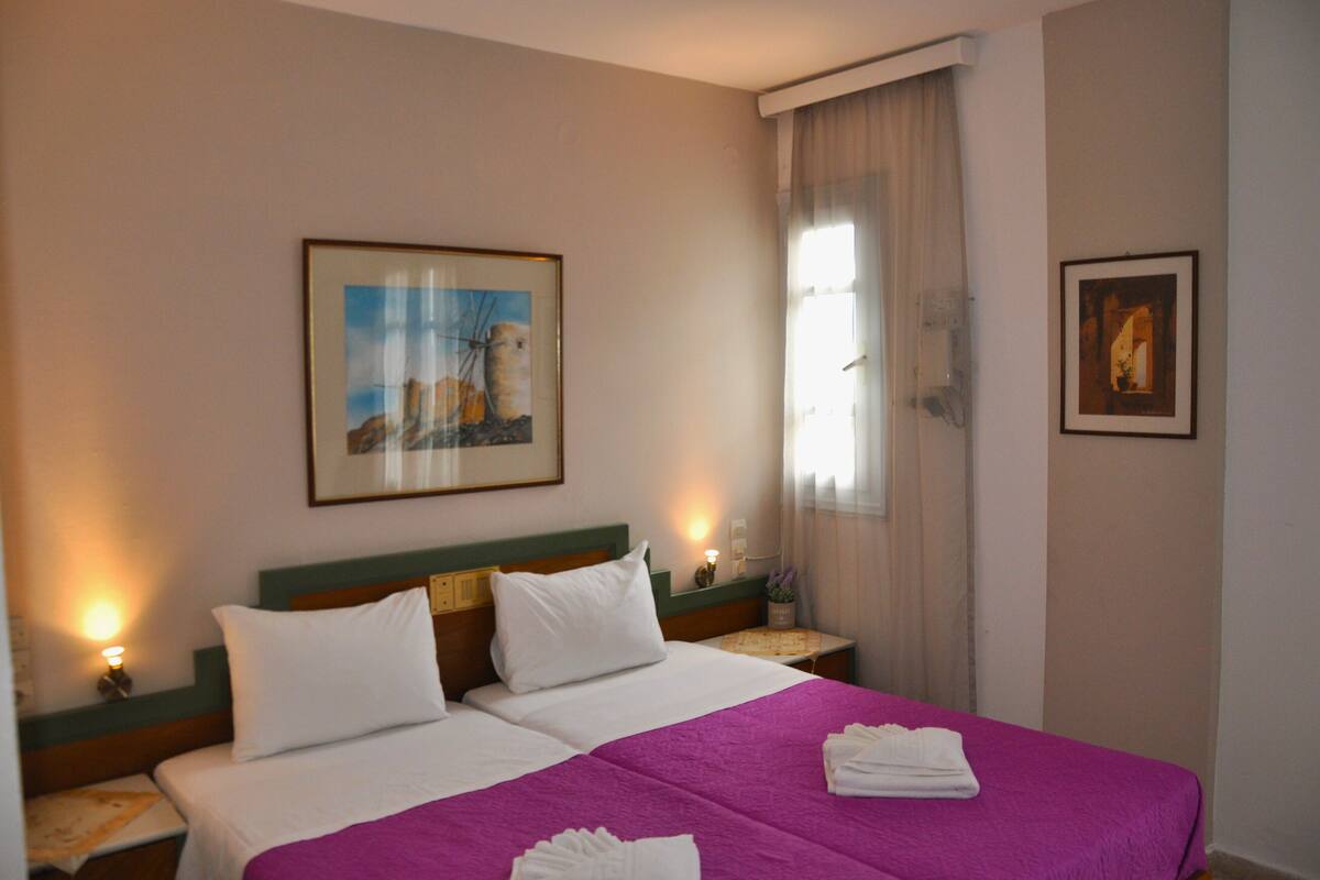 A hotel room with two beds, a purple bedspread, white pillows, and two framed pictures on the walls.