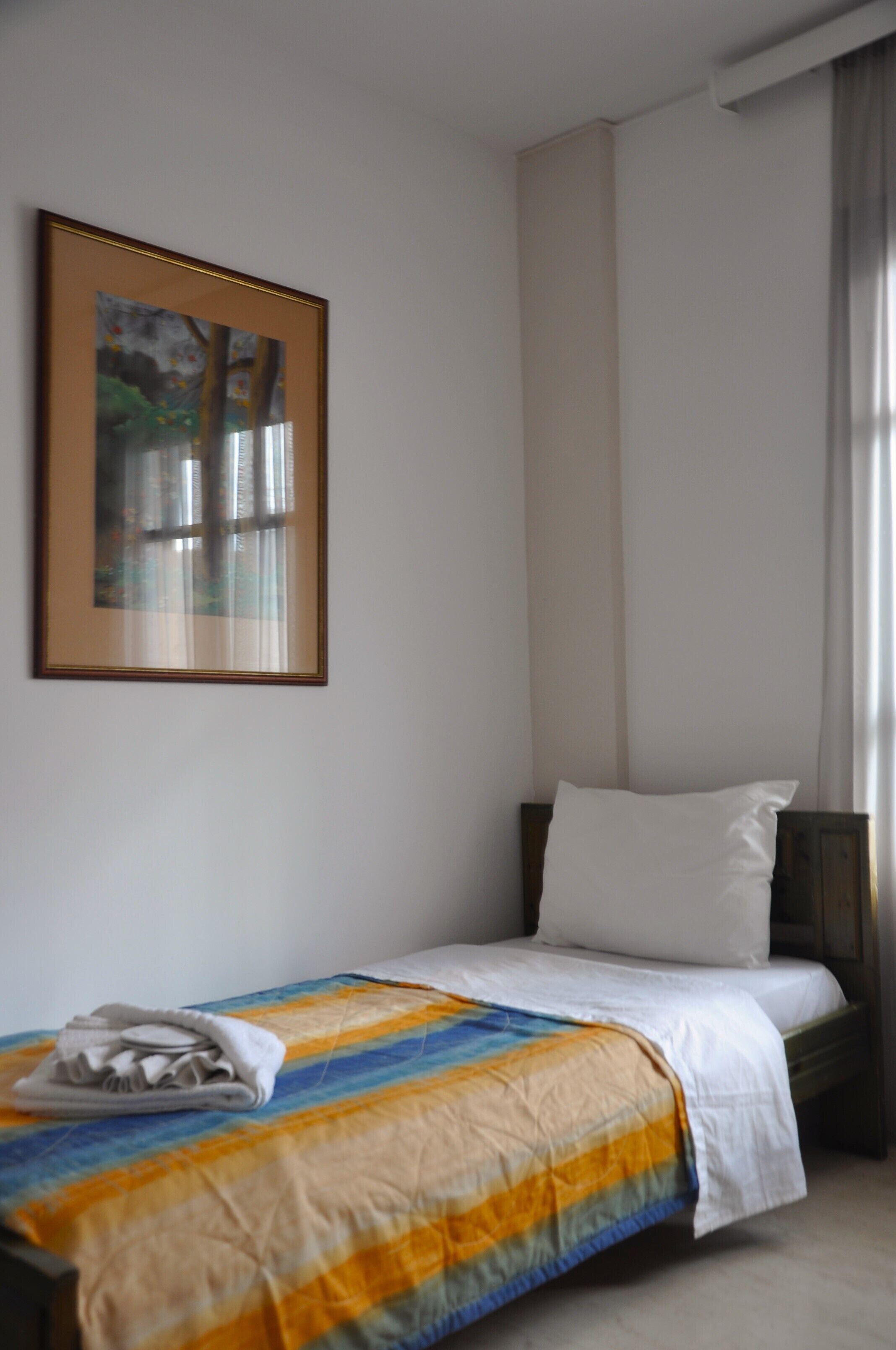 Una cama individual con una manta a rayas, una almohada blanca y una toalla doblada al lado. Un cuadro enmarco cuelga de la pared sobre la cama.
